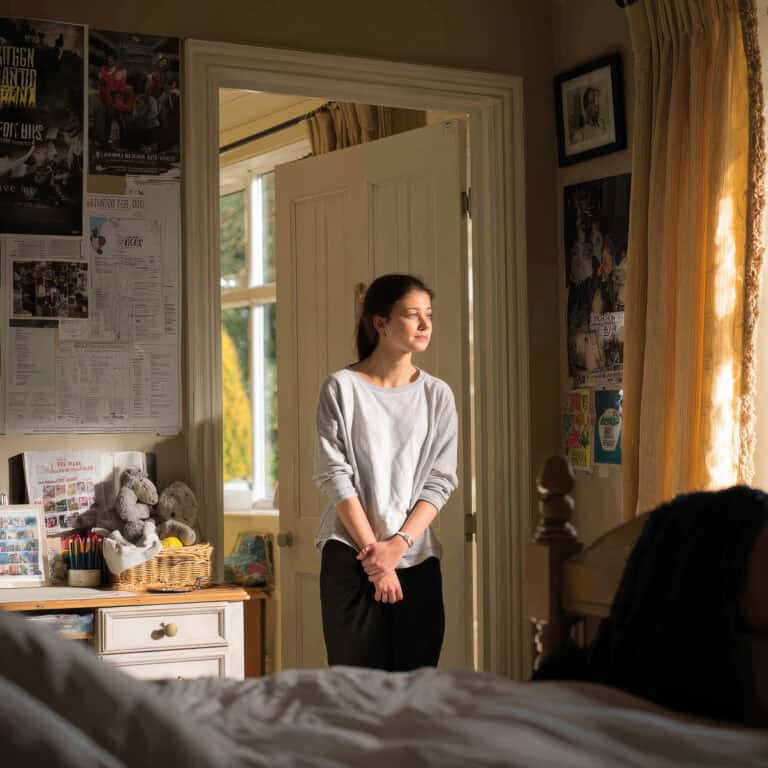 Listening to her thoughts, woman standing in a cozy, sunlit bedroom, surrounded by posters and personal items, reflecting relaxed yet thoughtful mood.
